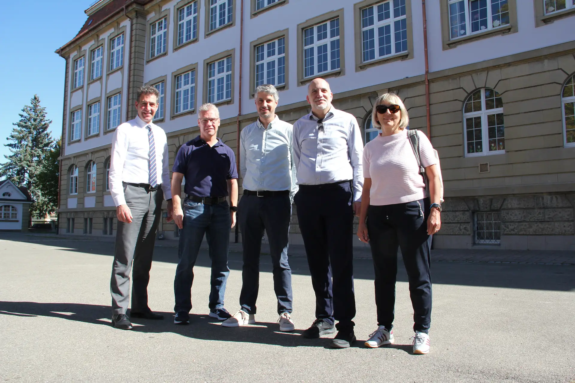 Personen stehen vor einem historischen Schulgebäude; im Vordergrund sind mehrere Männer und Frauen in formeller Kleidung zu sehen.